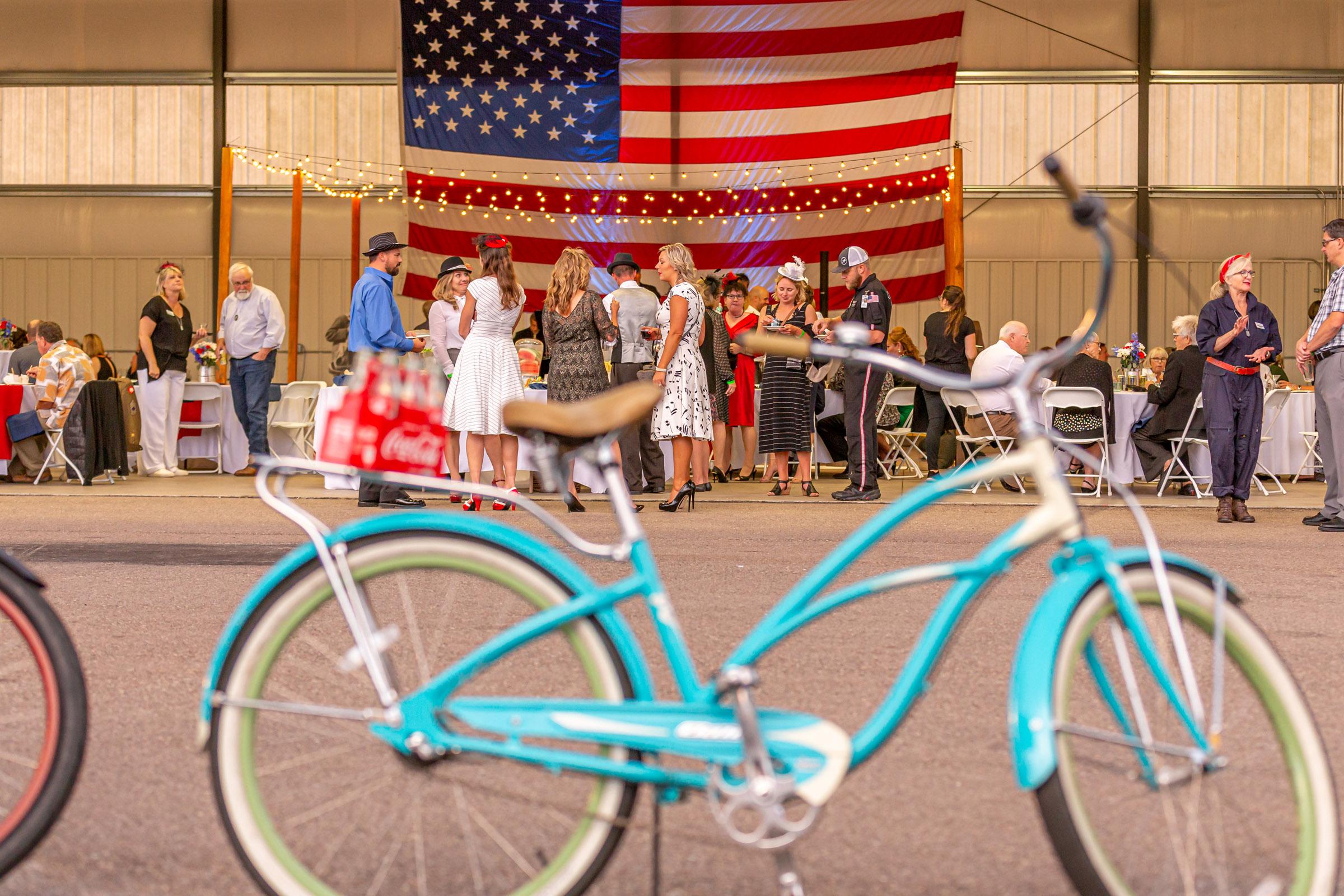 Hangar party guests under string lights and a large flag